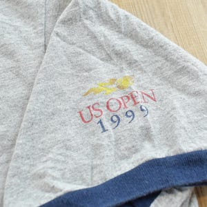 Puede incluir: Manga de camiseta gris con ribete azul marino. La manga muestra el texto "US OPEN 1999" en rojo y azul, con un logotipo amarillo y naranja encima. La tela tiene un aspecto ligeramente desgastado, lo que sugiere ropa vintage.