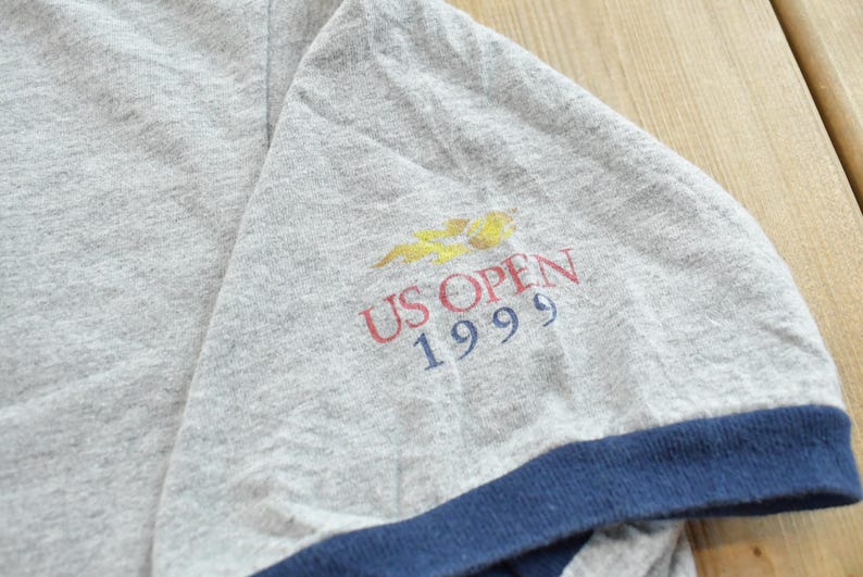 Puede incluir: Manga de camiseta gris con ribete azul marino. La manga muestra el texto "US OPEN 1999" en rojo y azul, con un logotipo amarillo y naranja encima. La tela tiene un aspecto ligeramente desgastado, lo que sugiere ropa vintage.