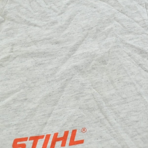 Op de afbeelding: Een lichtgrijs gem&ecirc;leerd t-shirt met het oranje STIHL-logo gedrukt nabij de onderzoom. De stof heeft een gespikkelde textuur over het gehele oppervlak en het kledingstuk bevat een geribbelde ronde hals in de linkerbovenhoek.