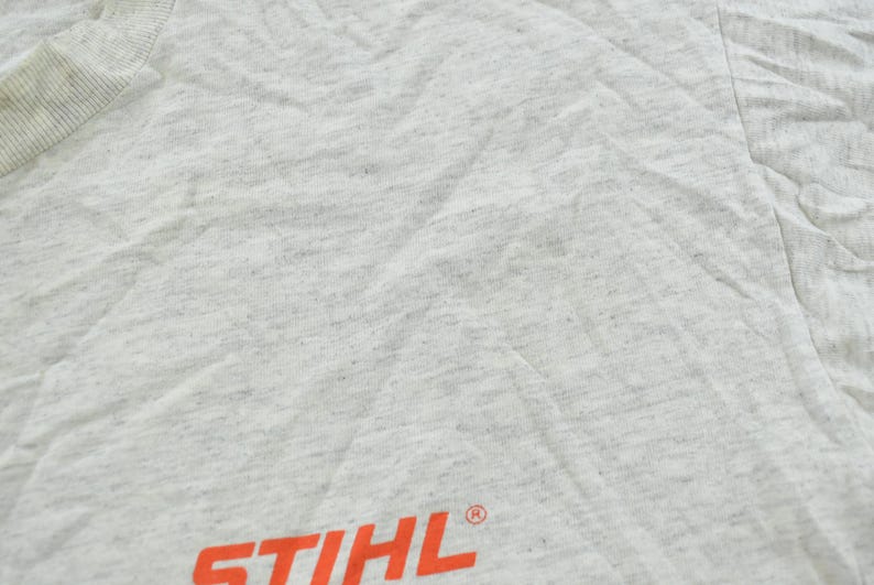Op de afbeelding: Een lichtgrijs gem&ecirc;leerd t-shirt met het oranje STIHL-logo gedrukt nabij de onderzoom. De stof heeft een gespikkelde textuur over het gehele oppervlak en het kledingstuk bevat een geribbelde ronde hals in de linkerbovenhoek.