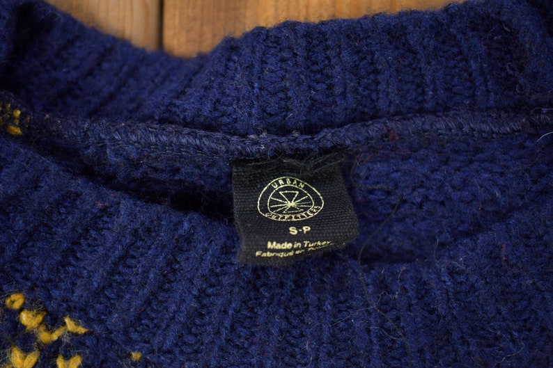 May include: A navy blue sweater with a yellow and white pattern. The sweater has a tag that says "Urban Outfitters" and "S-P" with the text "Made in Turkey" below.