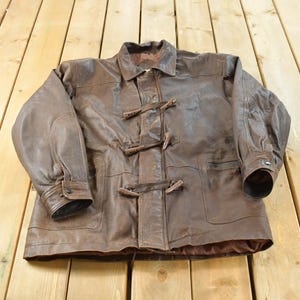 May include: A brown leather jacket featuring a classic collar, long sleeves with buttoned cuffs, and a front closure secured by three wooden toggle fasteners. The garment includes two large patch pockets on the front and a smooth brown interior lining.