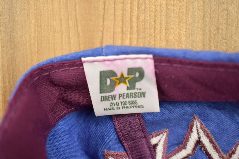May include: A blue and purple baseball cap with a white star and the text "D*P Drew Pearson (214) 702-8055 Made in Philippines" on a white tag.