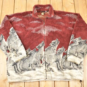 Peut inclure: Veste polaire bordeaux avec fermeture éclair intégrale. La veste présente un motif répété de loups hurlants en nuances de gris et de blanc sur un fond rouge. Les manches et le corps sont recouverts du motif loup.