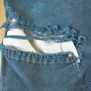 May include: Close-up of distressed blue denim jeans. The jeans have a large tear revealing a white fabric underneath. The pocket features a metal rivet. The denim has a worn, faded appearance.