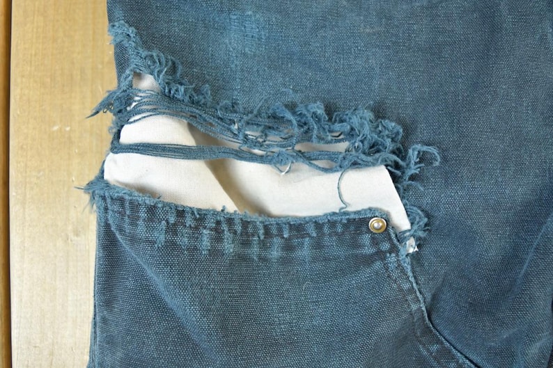 May include: Close-up of distressed blue denim jeans. The jeans have a large tear revealing a white fabric underneath. The pocket features a metal rivet. The denim has a worn, faded appearance.