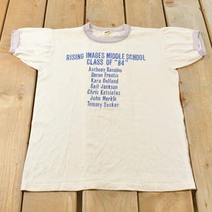 May include: A vintage white t-shirt with blue text. The text reads "RISING IMAGES MIDDLE SCHOOL CLASS OF '84" followed by a list of names. The shirt has light purple trim on the collar and sleeves. The shirt is laying on a wooden surface.