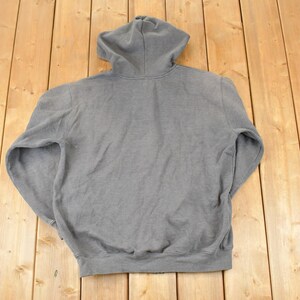 Puede incluir: Una sudadera con capucha gris con capucha con cord&oacute;n y mangas largas. La sudadera est&aacute; hecha de un material suave y texturizado. La prenda est&aacute; colocada sobre una superficie de madera.