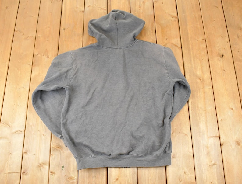 Puede incluir: Una sudadera con capucha gris con capucha con cord&oacute;n y mangas largas. La sudadera est&aacute; hecha de un material suave y texturizado. La prenda est&aacute; colocada sobre una superficie de madera.