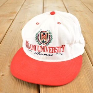 Może przedstawiać: Biało-czerwona czapka bejsbolowa z haftem "Miami University Alumni". Czapka ma czerwony daszek i logo z zielonym wieńcem i literą "M".