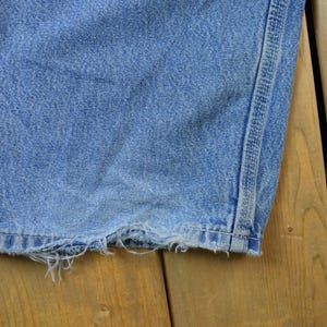 May include: Close-up of the bottom hem of a pair of blue denim jeans. The hem is frayed and has a single stitch along the edge.