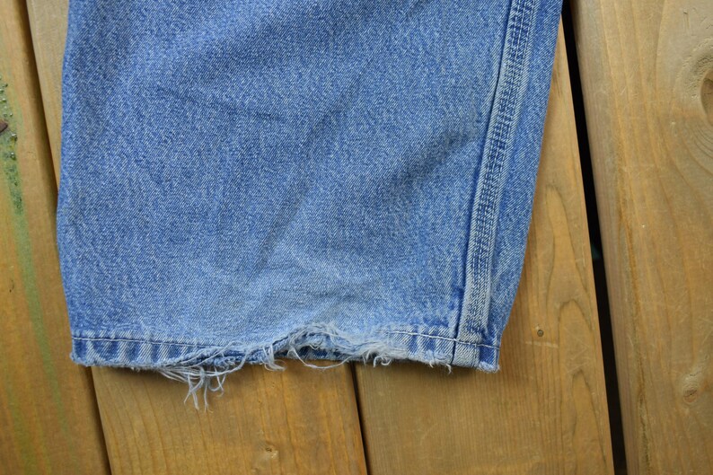 May include: Close-up of the bottom hem of a pair of blue denim jeans. The hem is frayed and has a single stitch along the edge.