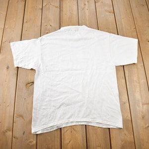 May include: A plain white t-shirt with short sleeves, laid flat on a wooden surface.