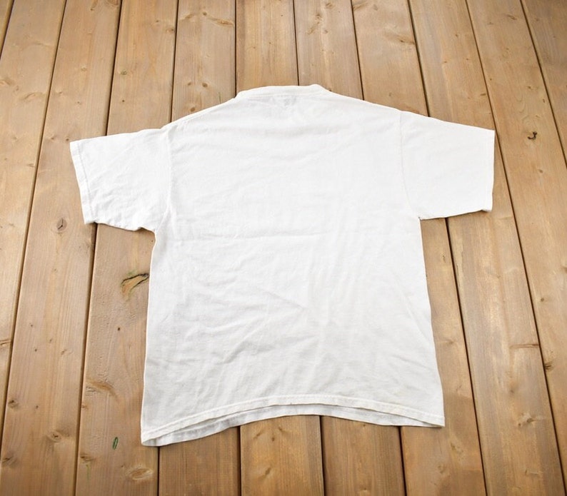 May include: A plain white t-shirt with short sleeves, laid flat on a wooden surface.