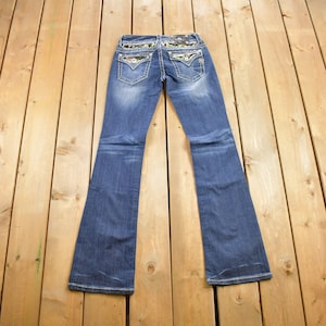 May include: A pair of blue denim jeans with a bootcut leg. The back pockets have a floral and camouflage print with rhinestones.
