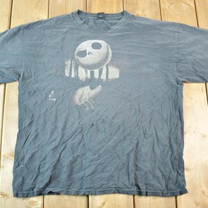 May include: A faded blue t-shirt featuring a graphic of Jack Skellington from The Nightmare Before Christmas. The design shows Jack's face and torso in a distressed, vintage style. The shirt is laid flat on a wooden surface.