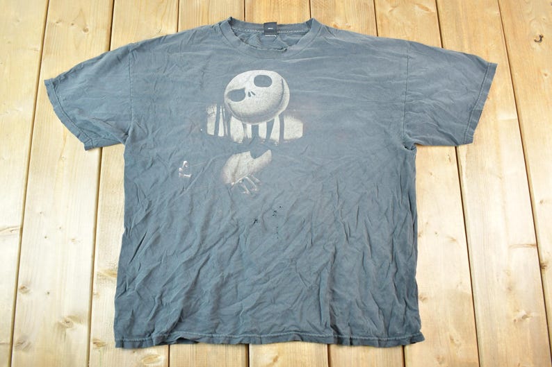 May include: A faded blue t-shirt featuring a graphic of Jack Skellington from The Nightmare Before Christmas. The design shows Jack's face and torso in a distressed, vintage style. The shirt is laid flat on a wooden surface.
