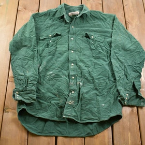 May include: A green long-sleeved button-down shirt with white paint splatters. The shirt has two chest pockets and a button-down collar.