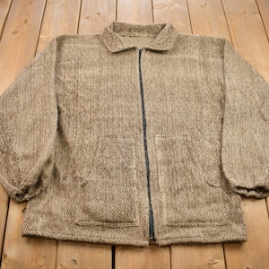 May include: A brown zippered jacket with a herringbone pattern. The jacket has two pockets on the front and a zip closure.