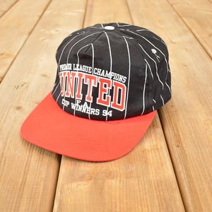Può includere: Cappellino da baseball nero e rosso con strisce verticali bianche. Il cappellino presenta la scritta "UNITED" a blocchi rossi, con "PREMIER LEAGUE CHAMPIONS" sopra e "CUP WINNERS 94" sotto.