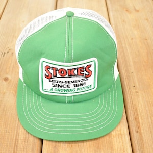 Può includere: Cappellino da camionista verde e bianco con visiera curva. Il cappello presenta un pannello frontale e una visiera verdi, un retro in rete bianca e una toppa con la scritta "Stokes Seeds-Semences Since 1881...A Growing Future."