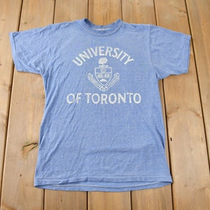 Puede incluir: Una camiseta azul claro con un gráfico blanco del escudo de la Universidad de Toronto y el texto "University of Toronto".