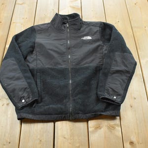 May include: A black full-zip jacket featuring a high collar, long sleeves, and a textured fleece lower body. The upper chest and sleeve panels are made of a smooth, solid black fabric. The North Face logo is embroidered in white on the chest.