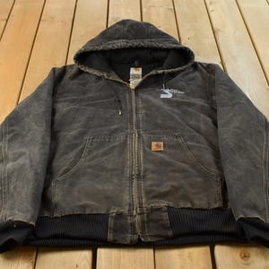May include: A charcoal grey hooded jacket with a full-length front zip, long sleeves, and ribbed cuffs and hem. The jacket features a small rectangular logo patch on the front pocket and a white graphic print on the upper chest area.