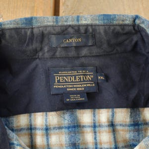 May include: Close-up of a Pendleton Canyon flannel shirt collar. The shirt features a blue, tan, and white plaid pattern. The collar is navy blue with a Canyon label. The Pendleton label is visible, indicating the brand and size XXL.