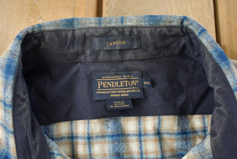 May include: Close-up of a Pendleton Canyon flannel shirt collar. The shirt features a blue, tan, and white plaid pattern. The collar is navy blue with a Canyon label. The Pendleton label is visible, indicating the brand and size XXL.