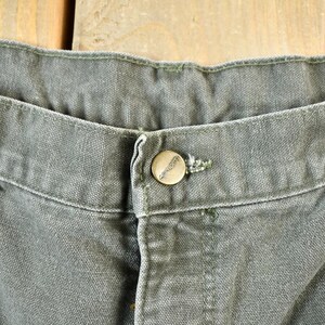 Pu&ograve; includere: Primo piano della cintura di un paio di pantaloni da lavoro grigi scuri con una singola chiusura a bottone.