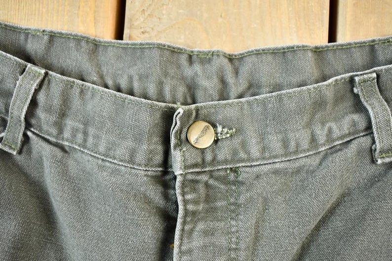 Pu&ograve; includere: Primo piano della cintura di un paio di pantaloni da lavoro grigi scuri con una singola chiusura a bottone.