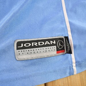 May include: A light blue athletic jersey with a white trim and a black and white Jordan Performance Brand of Excellence label.