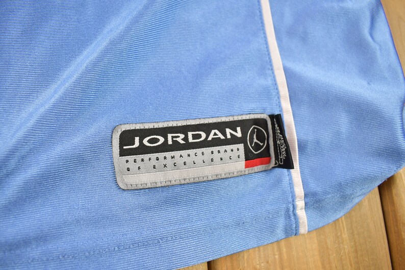 May include: A light blue athletic jersey with a white trim and a black and white Jordan Performance Brand of Excellence label.