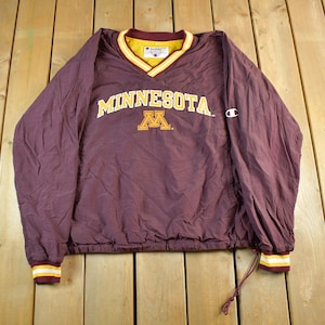 May include: A maroon Champion pullover jacket with a V-neck collar. The jacket features the word "MINNESOTA" in gold lettering across the chest, with a gold and maroon "M" logo below. The cuffs and collar have gold and white stripes.