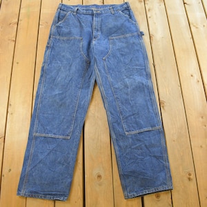 K&ouml;nnte beinhalten: Blaue Jeans-Arbeitshose mit verst&auml;rkten Kniepatches und mehreren Taschen. Die Hose liegt flach auf einer Holzoberfl&auml;che und zeigt ihre strapazierf&auml;hige Konstruktion und ihr klassisches Design. Ein zeitloses Kleidungsst&uuml;ck.