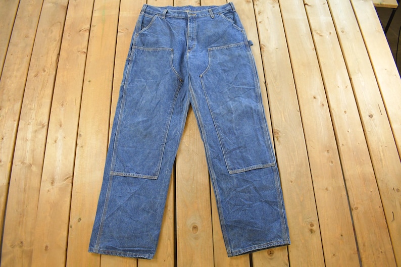 K&ouml;nnte beinhalten: Blaue Jeans-Arbeitshose mit verst&auml;rkten Kniepatches und mehreren Taschen. Die Hose liegt flach auf einer Holzoberfl&auml;che und zeigt ihre strapazierf&auml;hige Konstruktion und ihr klassisches Design. Ein zeitloses Kleidungsst&uuml;ck.