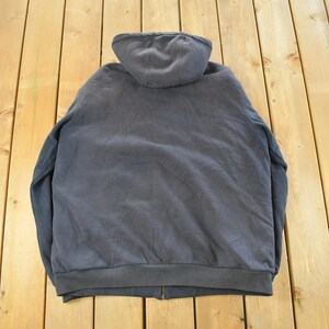 Puede incluir: Una sudadera con capucha gris oscuro con cierre de cremallera completo. Se ve la parte trasera de la sudadera, mostrando la capucha y el dobladillo inferior. La prenda es de material suave.