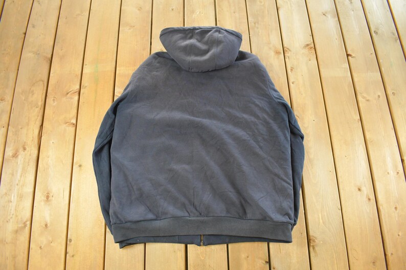 Puede incluir: Una sudadera con capucha gris oscuro con cierre de cremallera completo. Se ve la parte trasera de la sudadera, mostrando la capucha y el dobladillo inferior. La prenda es de material suave.