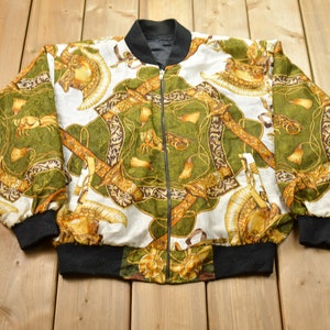 May include: A white bomber jacket with a black zipper and black ribbed cuffs and waistband. The jacket has a green and gold patterned design featuring swords, chains, and other ornate details.