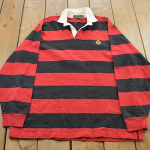 May include: Red and black striped long-sleeve rugby shirt with a white collar. The shirt features a small gold emblem on the chest. The shirt is made of a soft, comfortable material.