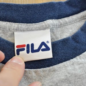 Puede incluir: Primer plano de una camiseta gris con cuello y ribetes de manga azul marino. La camiseta presenta una etiqueta blanca con el logotipo de FILA en azul marino y rojo.