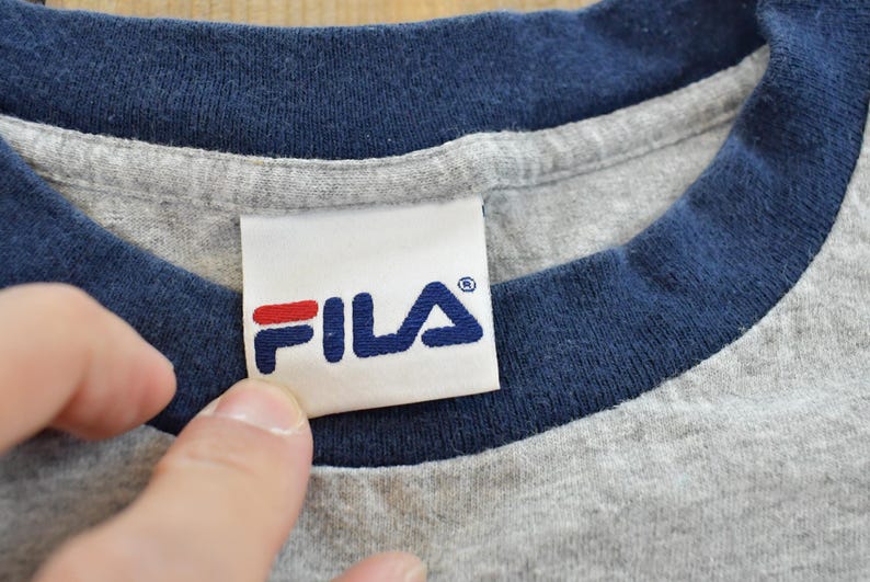 Puede incluir: Primer plano de una camiseta gris con cuello y ribetes de manga azul marino. La camiseta presenta una etiqueta blanca con el logotipo de FILA en azul marino y rojo.
