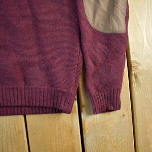 Puede incluir: Primer plano de un su&eacute;ter burdeos con dobladillo y pu&ntilde;os acanalados. El su&eacute;ter tiene un parche marr&oacute;n en el codo. La prenda de punto est&aacute; hecha de un material texturizado. El fondo es una superficie de madera marr&oacute;n claro.