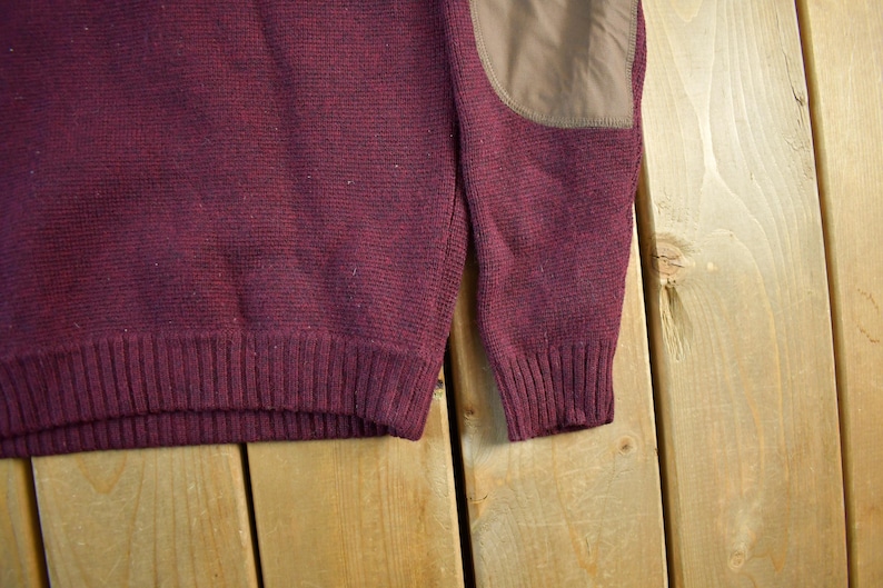 Puede incluir: Primer plano de un su&eacute;ter burdeos con dobladillo y pu&ntilde;os acanalados. El su&eacute;ter tiene un parche marr&oacute;n en el codo. La prenda de punto est&aacute; hecha de un material texturizado. El fondo es una superficie de madera marr&oacute;n claro.