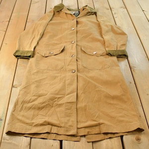 May include: A tan long-length coat featuring a contrasting olive green corduroy collar and matching sleeve cuffs. The coat includes a front button-down closure with metallic buttons and two large patch pockets on the front with buttoned flaps.