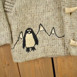 May include: Close-up of a light gray knit sweater with a penguin and mountain design. The sweater has a textured appearance and wooden toggle closures. The penguin is black and cream-colored, with a mountain range outline.
