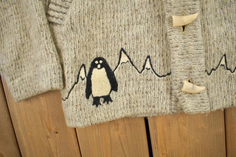 May include: Close-up of a light gray knit sweater with a penguin and mountain design. The sweater has a textured appearance and wooden toggle closures. The penguin is black and cream-colored, with a mountain range outline.