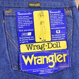Op de afbeelding: Een blauw denim jean-label met de tekst "Wrag-Doll" en "Wrangler" in geel. Het label bevat ook de tekst "Guarantee" en "Garantie" in het Engels en Frans, samen met details over de denim en de maat.