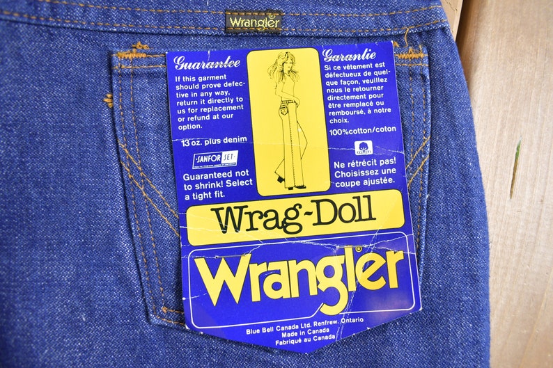 Op de afbeelding: Een blauw denim jean-label met de tekst "Wrag-Doll" en "Wrangler" in geel. Het label bevat ook de tekst "Guarantee" en "Garantie" in het Engels en Frans, samen met details over de denim en de maat.